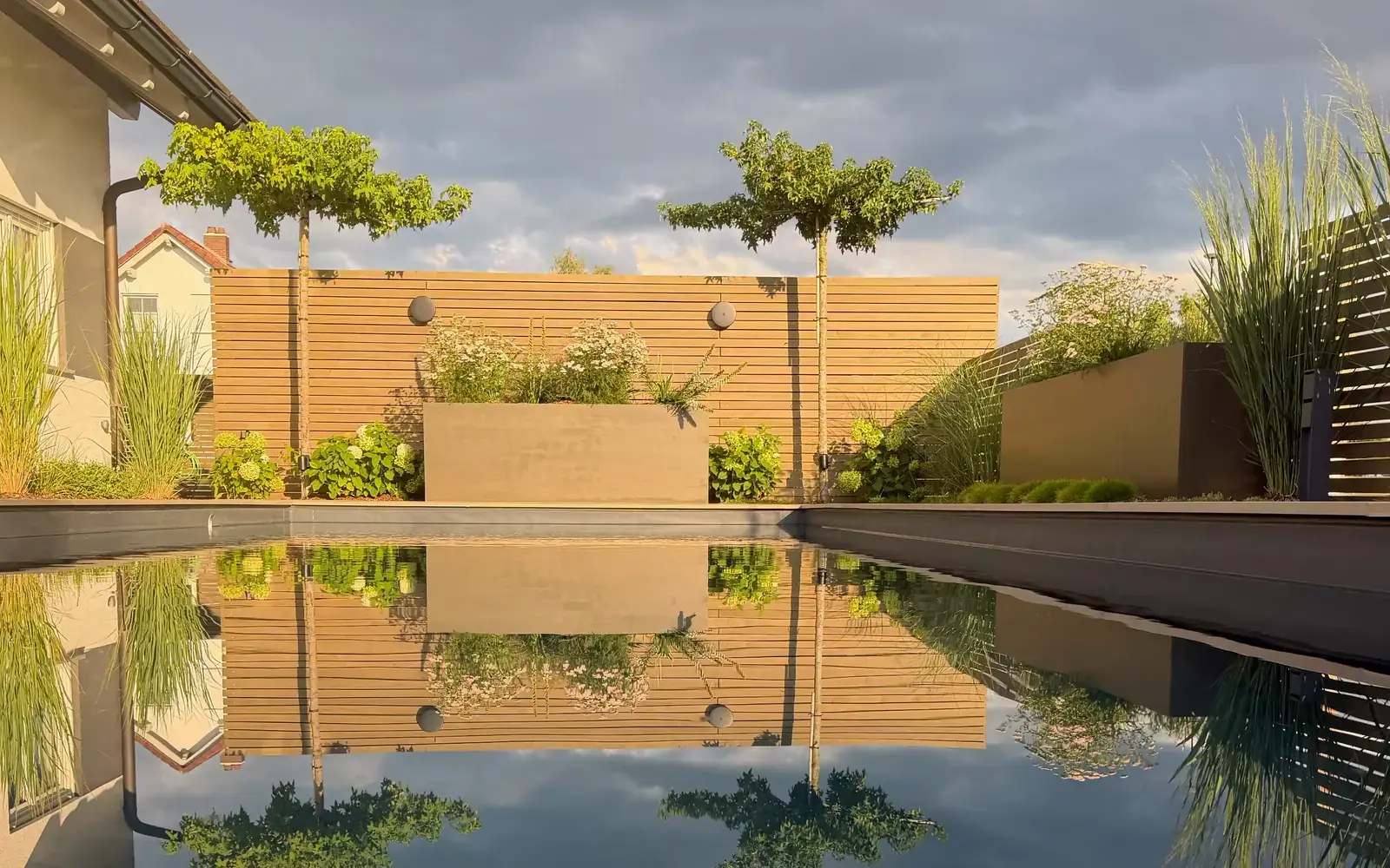 Pool im Abendlicht, in dem sich eine Holzwand mit Hochbeet, zwei Bäume und Ziergräser spiegeln