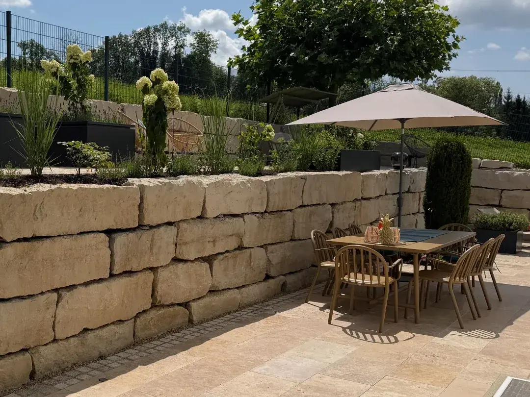 Nachher: Terrasse aus hellem Naturstein vor einer Natursteinmauer mit Staudenbeet, Hortensien und Ziergräsern, dazu Esstisch mit Stühlen unter einem Sonnenschirm