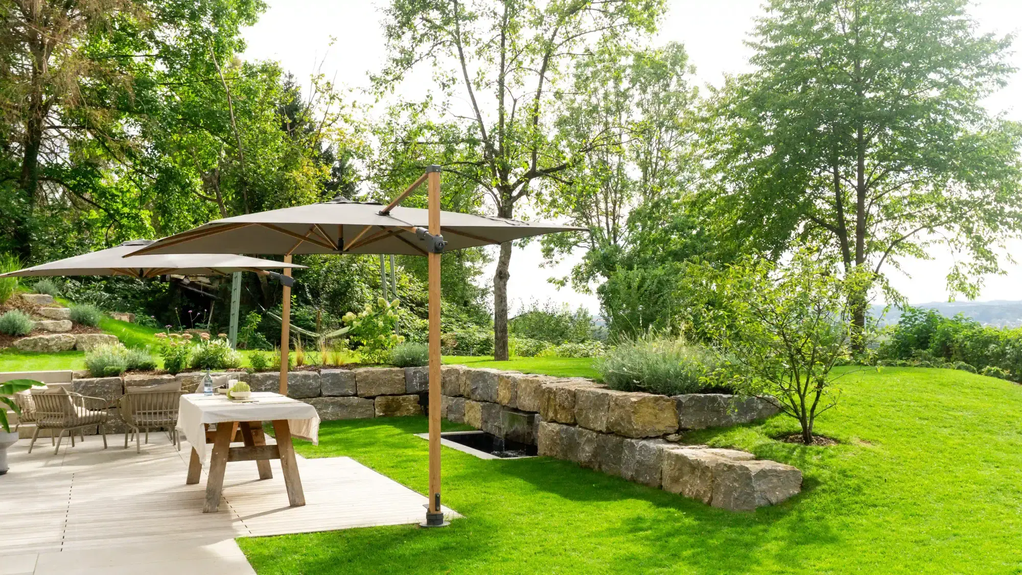 Gartenbau: Natursteinmauer mit Wasserlauf, gepflegter Rasen und Terrasse mit Holztisch, Stühlen und zwei Sonnenschirmen vor hohen Bäumen