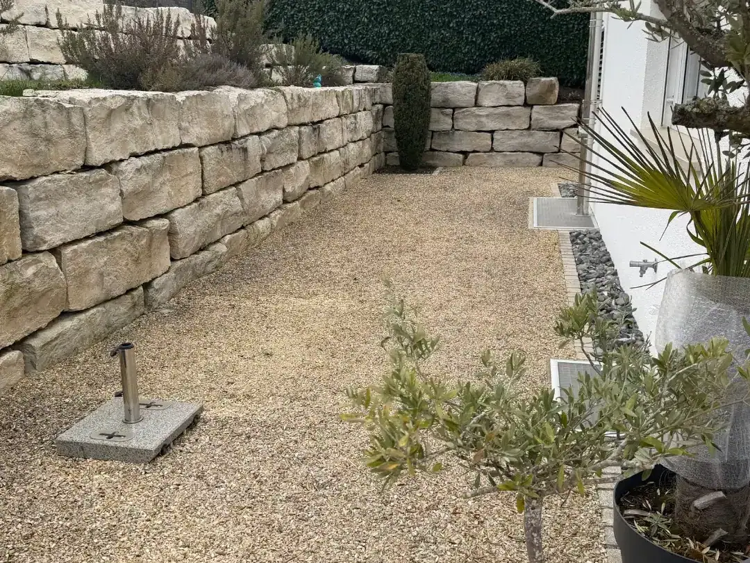 Vorher: Kiesfläche neben einem Haus, links eine Natursteinmauer aus großen Kalksteinblöcken, davor ein Schirmständer und ein Olivenbaum im Topf