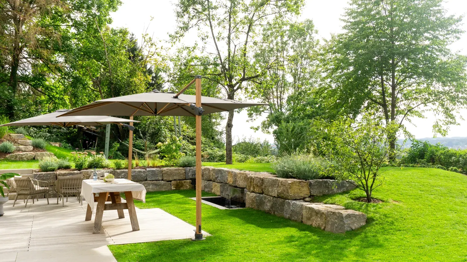 Terrasse mit Esstisch und Stühlen unter großen Sonnenschirmen, umgeben von Bäumen und einer Natursteinmauer.
