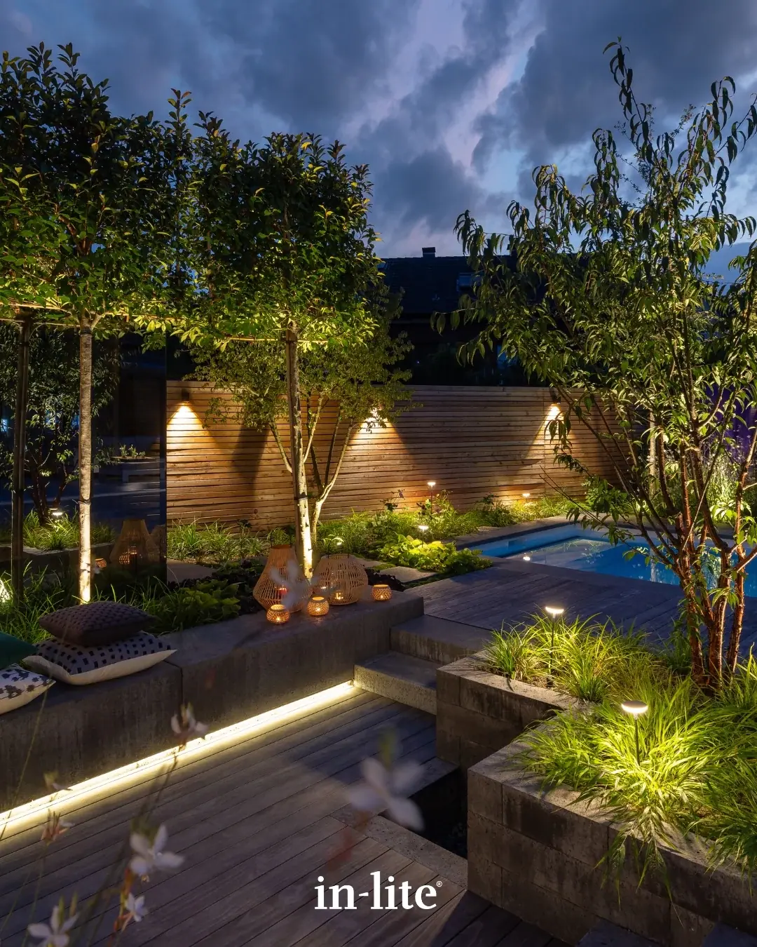 Abendlicher Garten mit beleuchteten Bäumen, Pflanzbeeten und Pool.