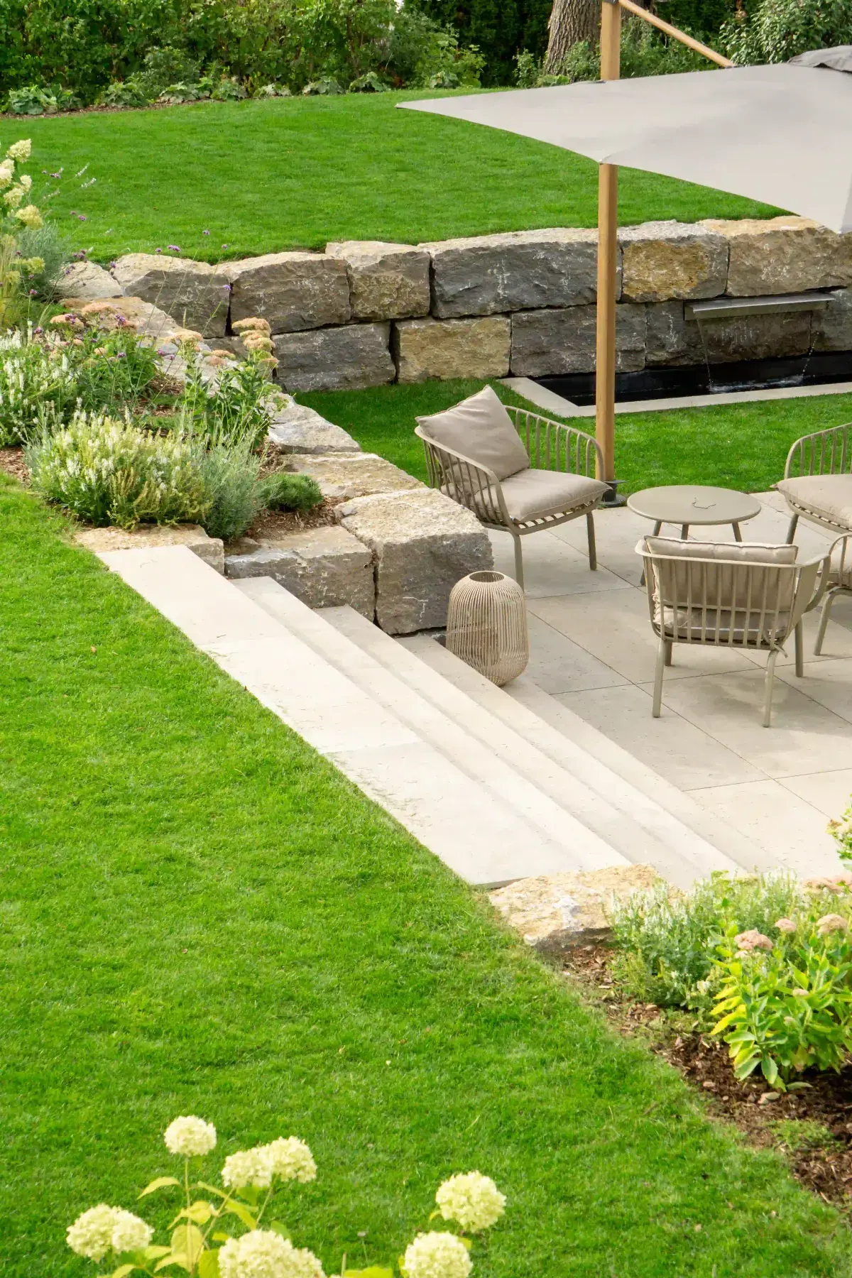 Sitzplatz mit Loungesesseln und Sonnensegel neben Natursteinmauer, Wasserlauf, Stufen und Staudenbeet in einem individuell geplanten Garten