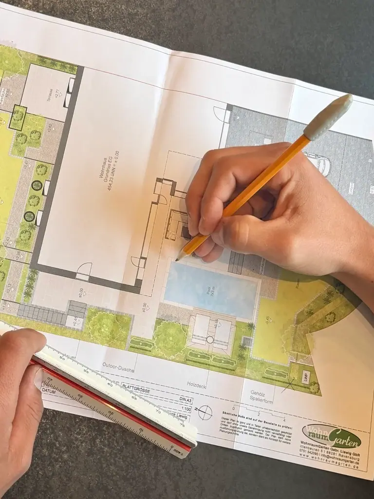 Hände arbeiten mit Bleistift und Lineal an einem farbigen Gartenplan mit Pool, Terrasse und Bepflanzung.