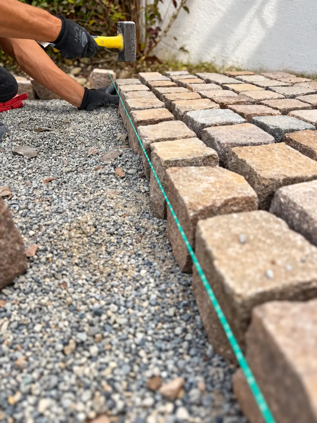 Hände mit Arbeitshandschuhen richten Pflastersteine mit einem Hammer entlang einer gespannten Schnur aus.