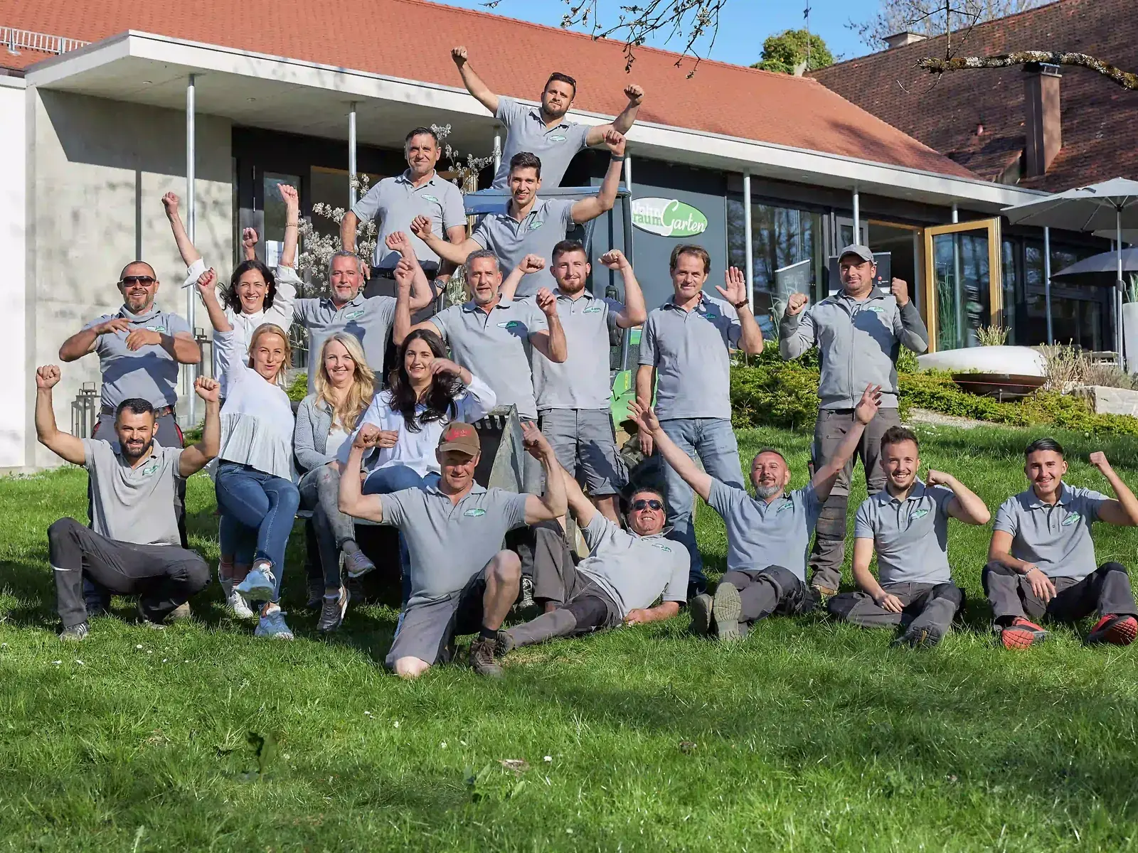 Das Team von WohnraumGarten jubelt gemeinsam auf einer Wiese vor dem Firmengebäude, die meisten in grauen Poloshirts mit Firmenlogo, einige sitzend und kniend vorn