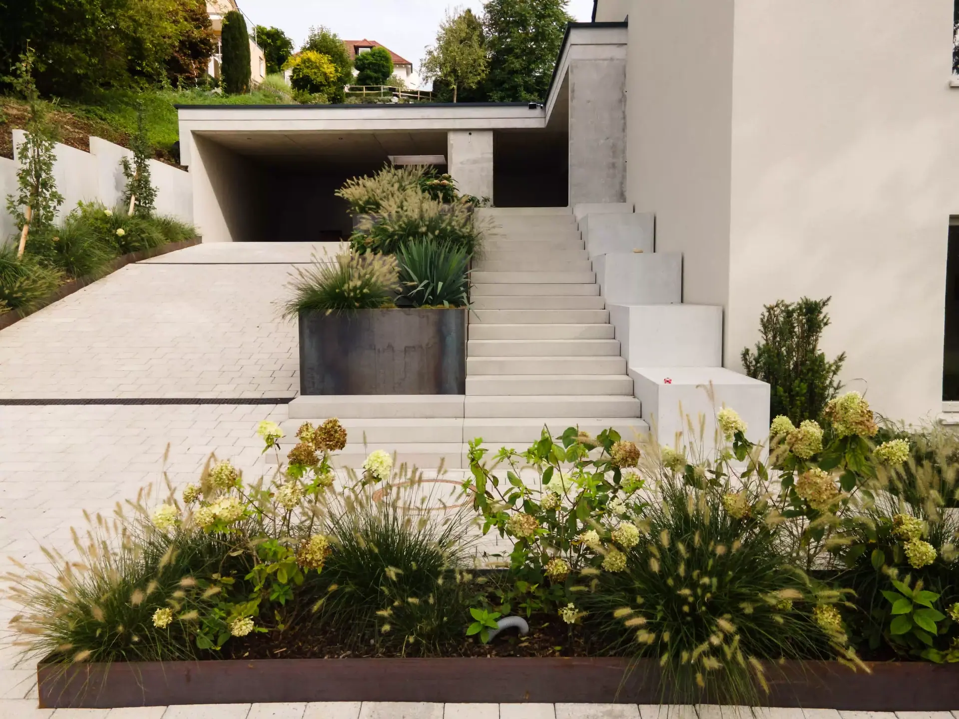 Einfahrt mit heller Treppe und Pflanzbeeten vor einem Wohnhaus.