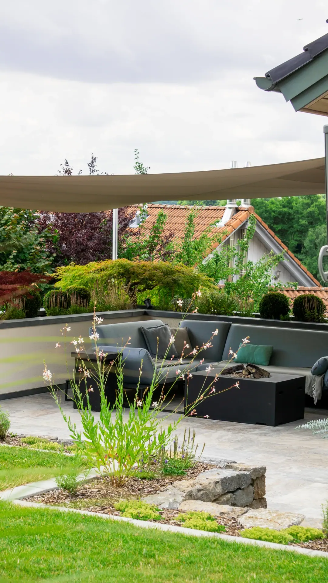 Gartenlounge unter einem Sonnensegel mit Ziergehölzen.