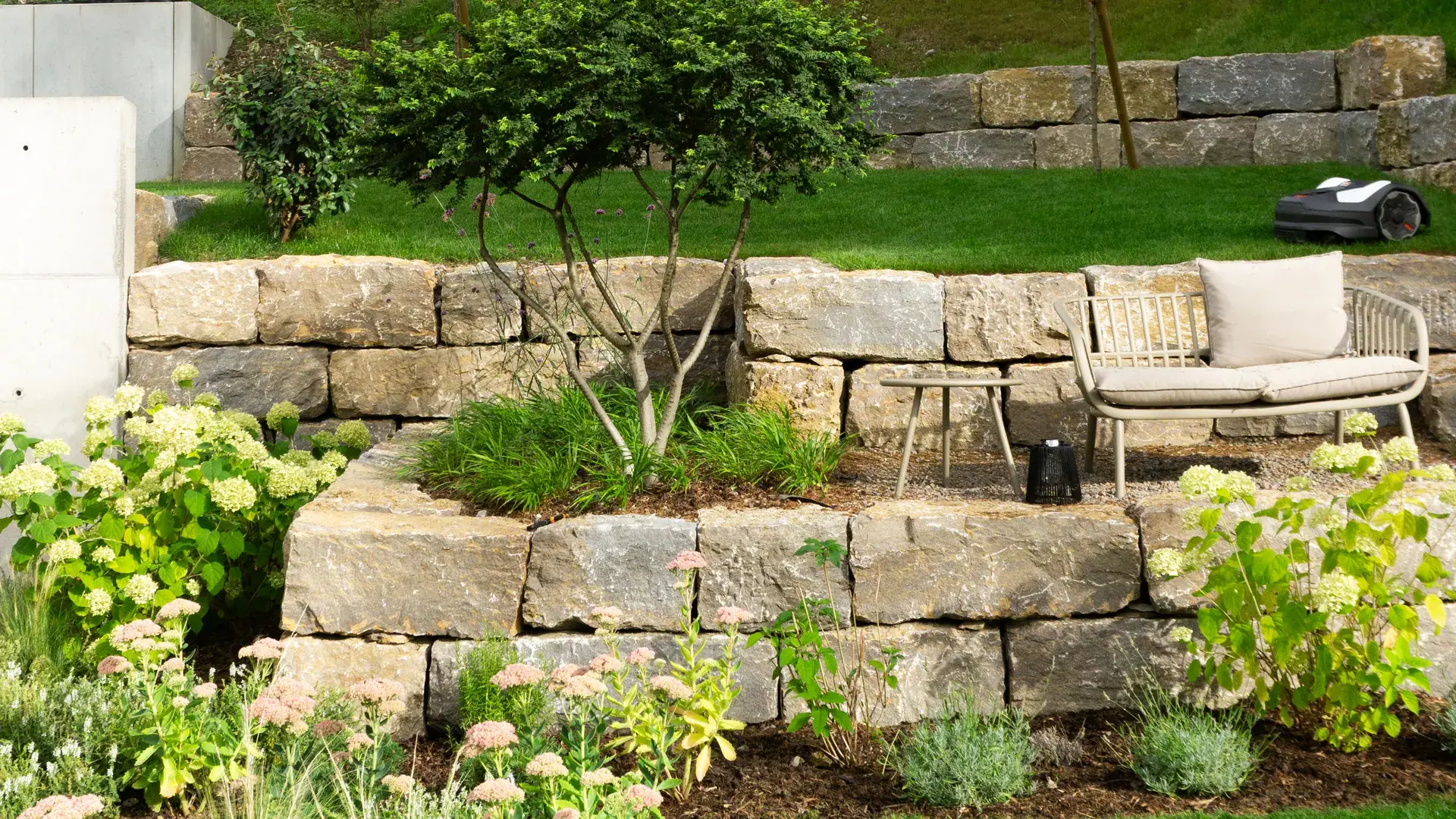 Natursteinmauer mit Sitzbank und blühenden Beeten.