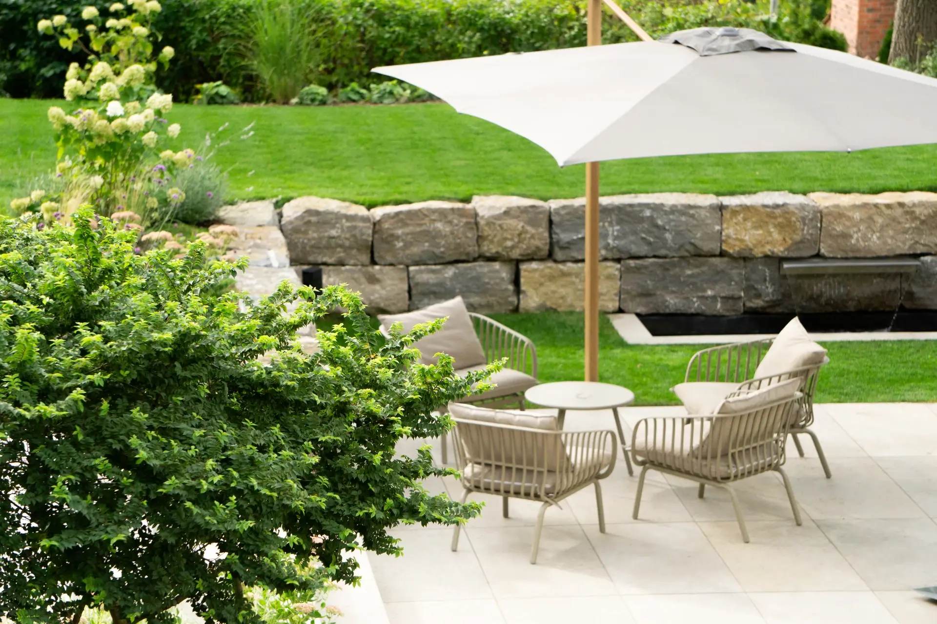 Sitzgruppe mit Sonnenschirm vor Natursteinmauer und Blumenbeet.