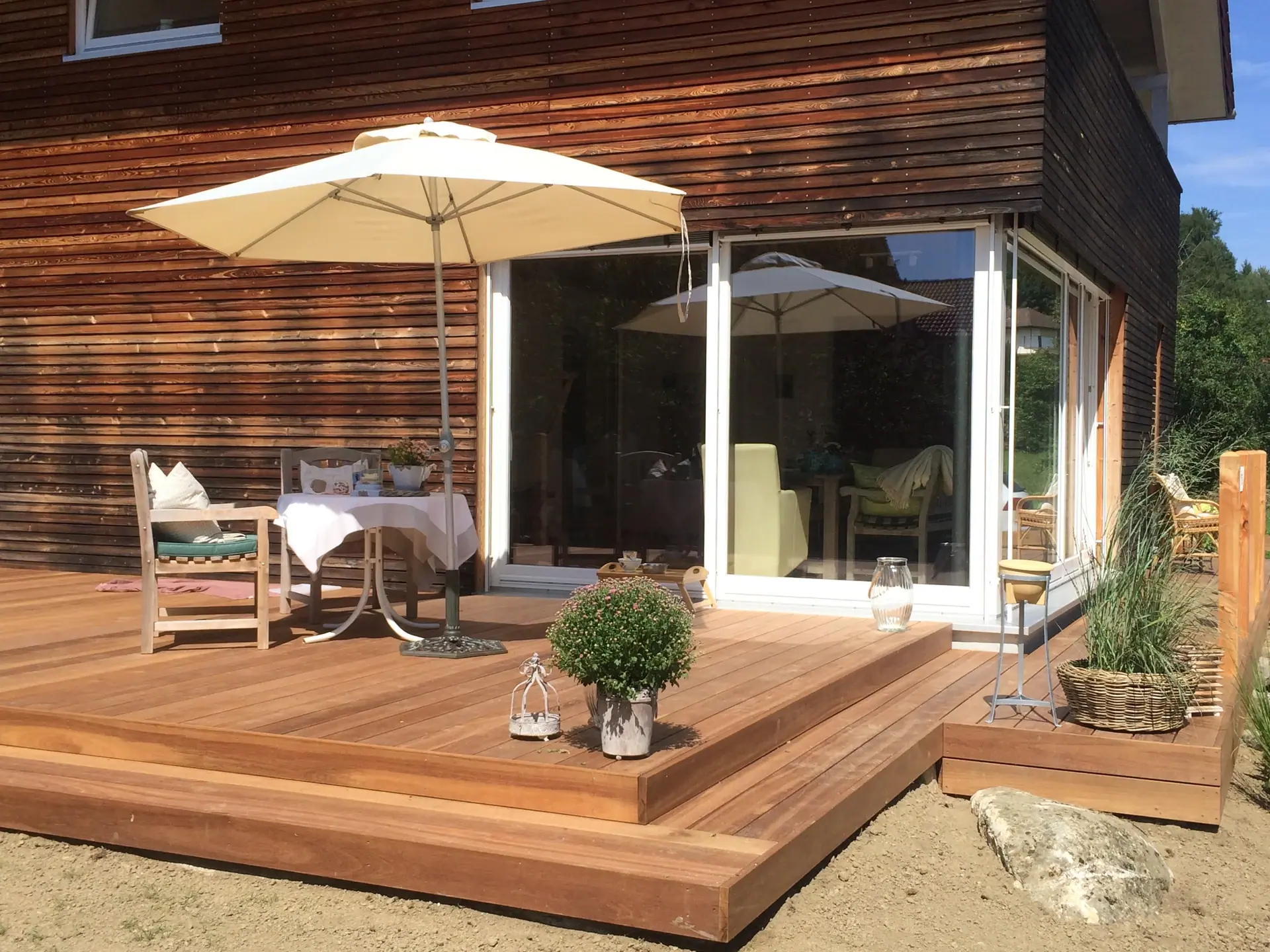 Holzterrasse mit Gartentisch und Sonnenschirm vor einem Haus.
