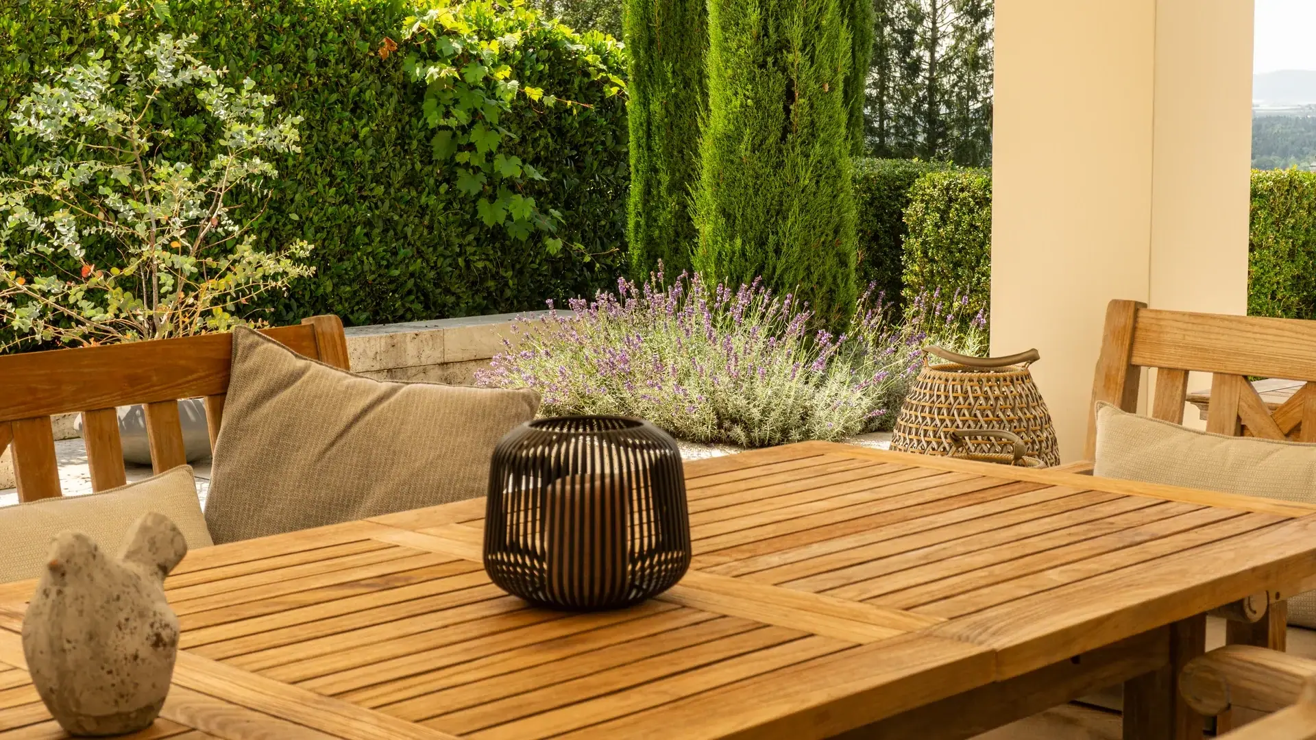 Holztisch mit Kissen und Sitzbank auf einer begrünten Terrasse.