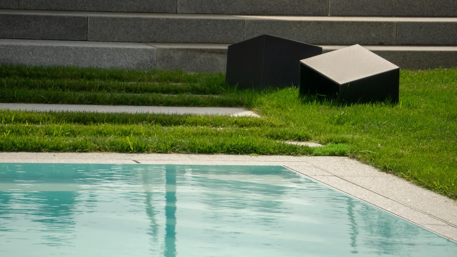 Poolrand neben Rasen und flachen schwarzen Einbauten.