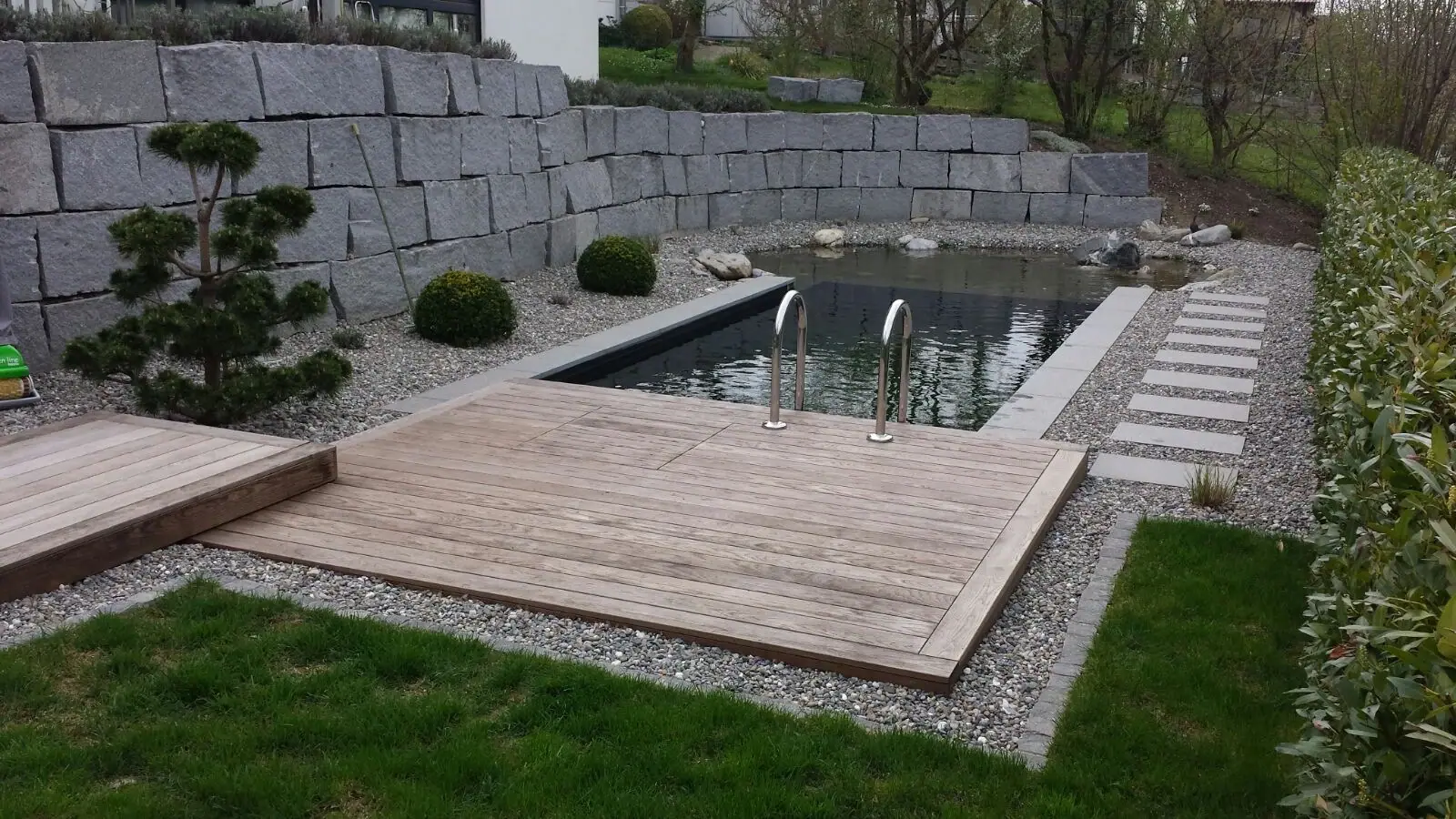 Schwimmteich mit Holzdeck vor einer Natursteinmauer.