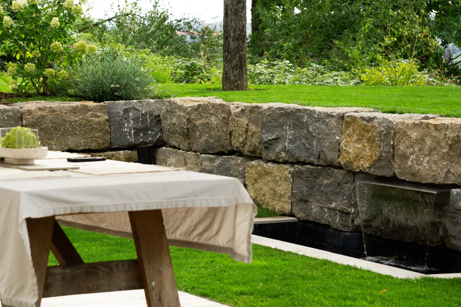 Sitzplatz vor einer Natursteinmauer mit Wasserlauf.
