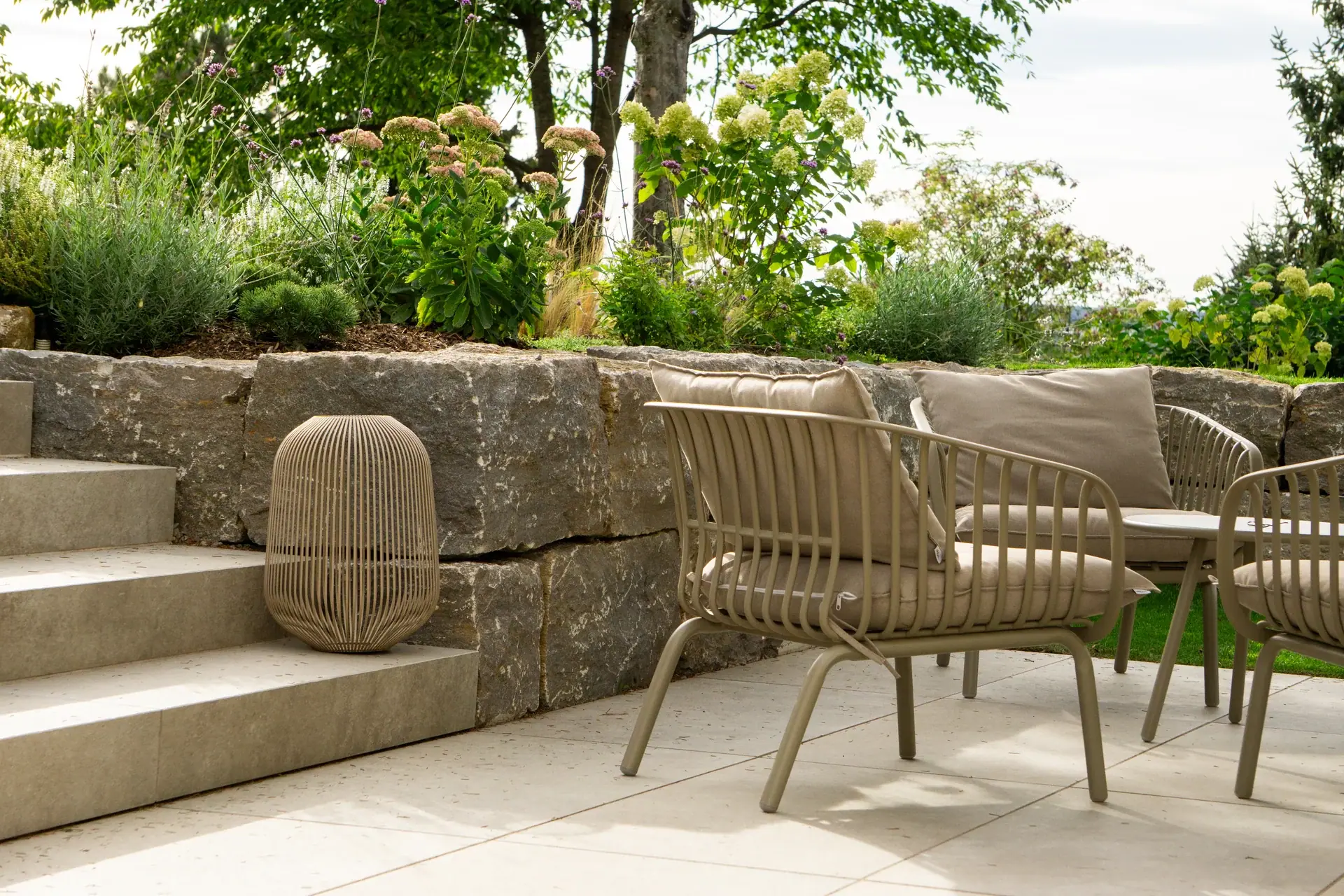 Terrasse mit hellen Gartenstühlen vor einer Natursteinmauer.