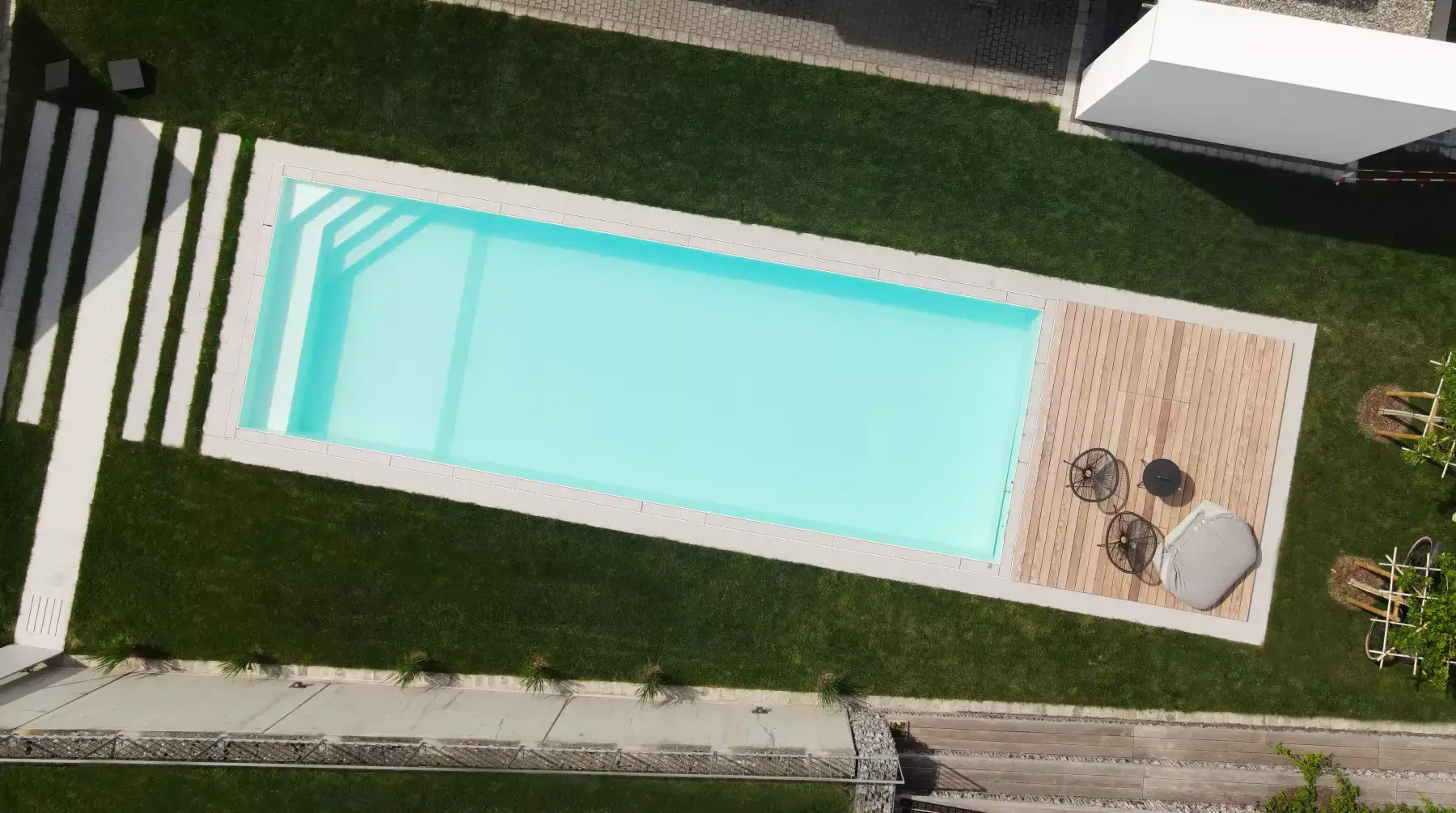 Luftbild eines Gartens mit rechteckigem Pool in Granitumrandung, Holzdeck mit Sitzgelegenheiten, Rasenflächen und Trittplatten