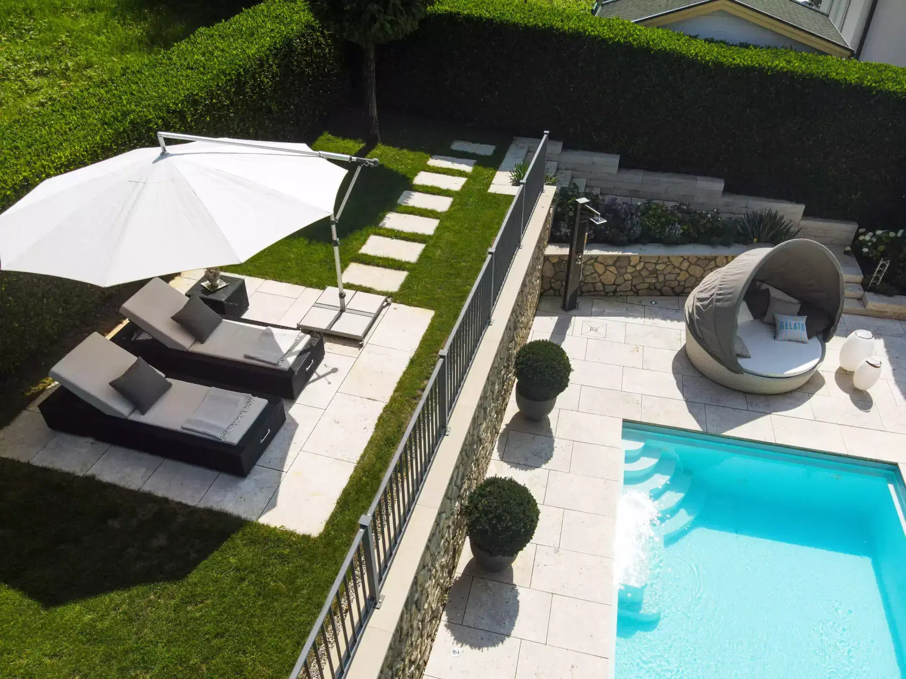 Blick von oben auf einen Garten mit Pool, hellen Natursteinplatten, Daybed, zwei Sonnenliegen unter einem Schirm und einer hohen Hecke
