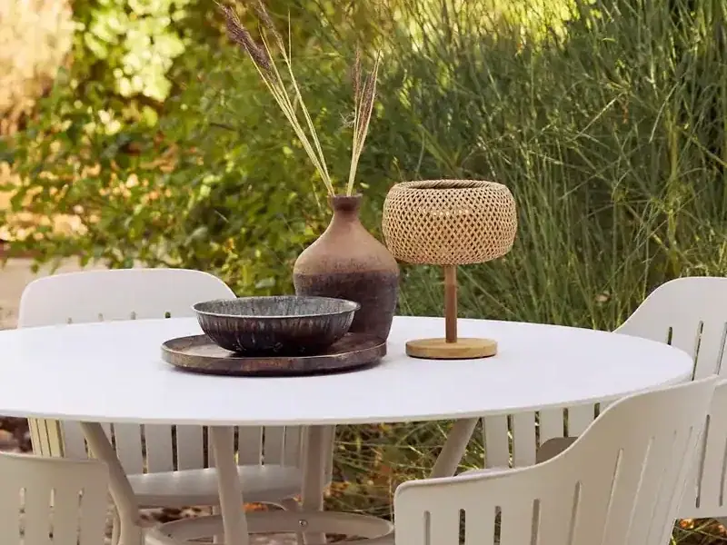 Rattan-Tischleuchte, Keramikvase mit Gräsern und Metallschale als Dekoration auf einem runden weißen Esstisch im Garten.