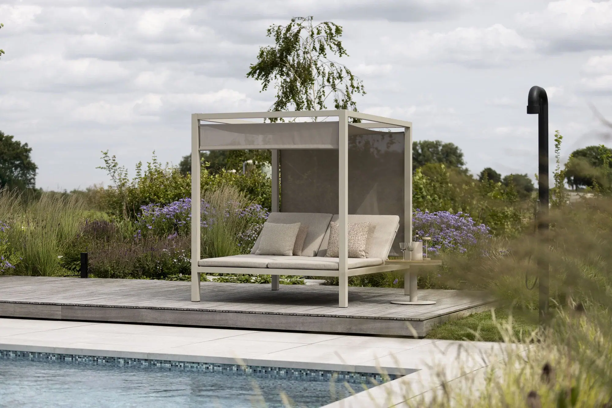 Daybed mit Sonnendach, hellen Polstern und kleinem Beistelltisch auf einem Holzdeck am Pool, umgeben von blühenden Stauden und Gräsern.