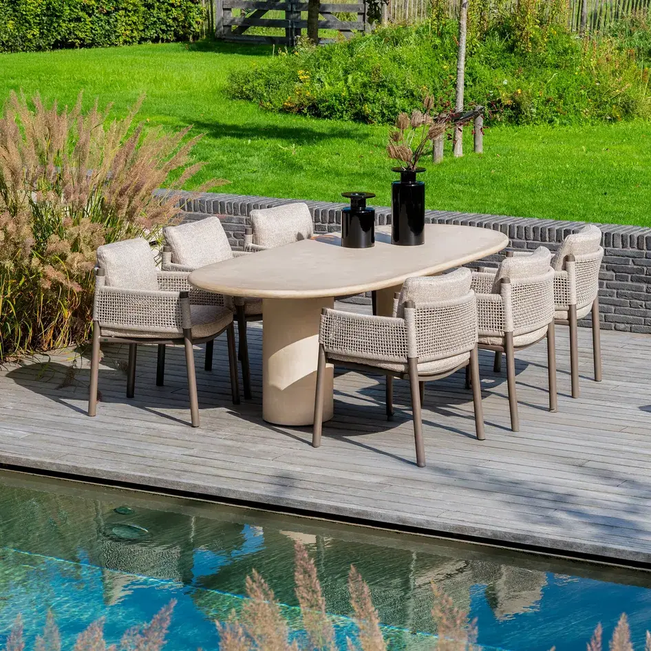Ovaler Esstisch mit hellem Sockel und sechs Geflechtstühlen auf einer Holzterrasse am Pool, dahinter Rasen und eine Backsteinmauer.