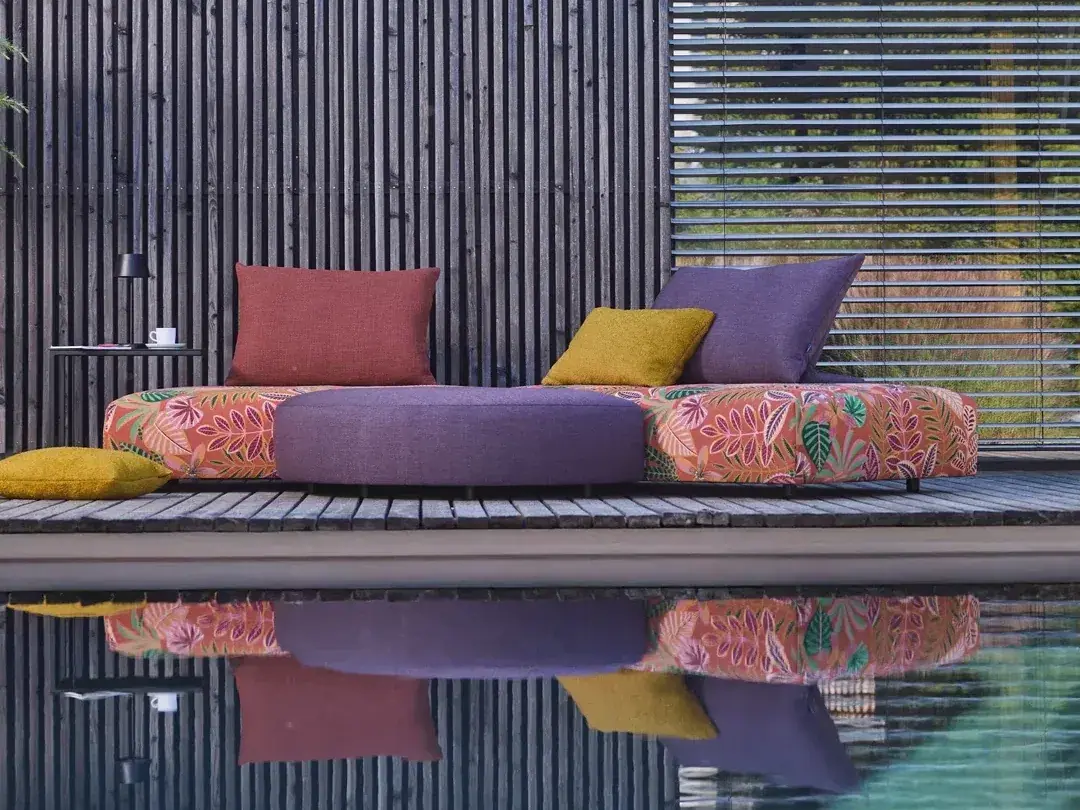 Farbenfrohes Outdoor-Sofa mit Blumenmuster und bunten Kissen in Rot, Gelb und Violett auf einer Holzterrasse am Pool.