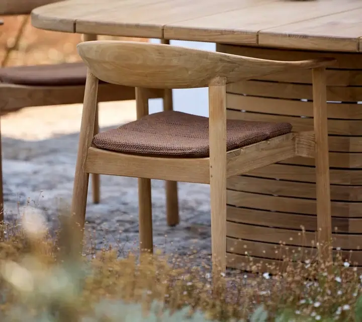 Gartenstuhl aus Teakholz mit braunem Sitzkissen an einem runden Esstisch aus Teakholz auf einer Terrasse.