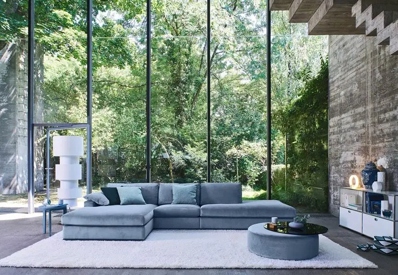 Modernes Wohnzimmer mit hellblauem Sofa, rundem Pouf und weißem Hochflorteppich vor einer bodentiefen Glasfront mit Blick in den Wald.