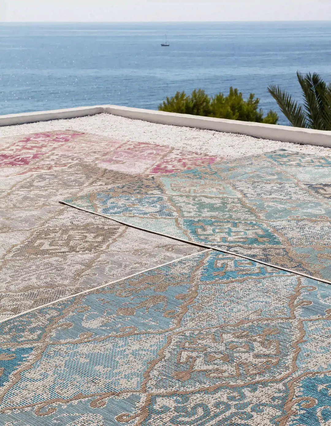 Mehrere gemusterte Outdoor-Teppiche in Blau-, Rosa- und Grautönen auf einer Terrasse mit Blick aufs Meer.