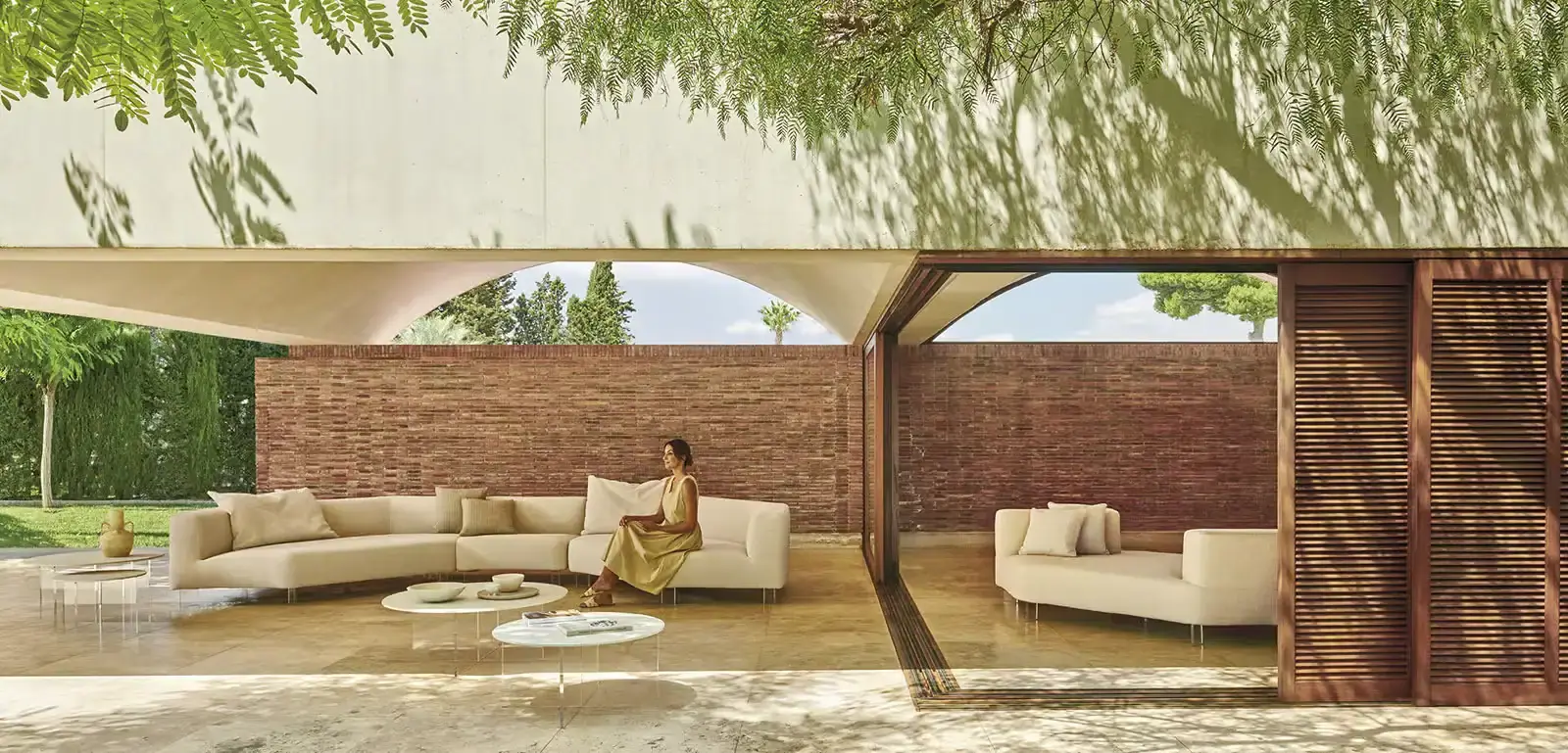 Cremefarbenes Loungesofa mit Beistelltischen auf einer überdachten Terrasse mit Backsteinwand, eine Frau sitzt darauf.