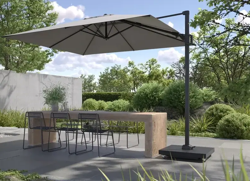Grauer Ampelschirm über einem Holztisch mit schwarzen Stühlen auf einer Terrasse zwischen Gräsern und Büschen.