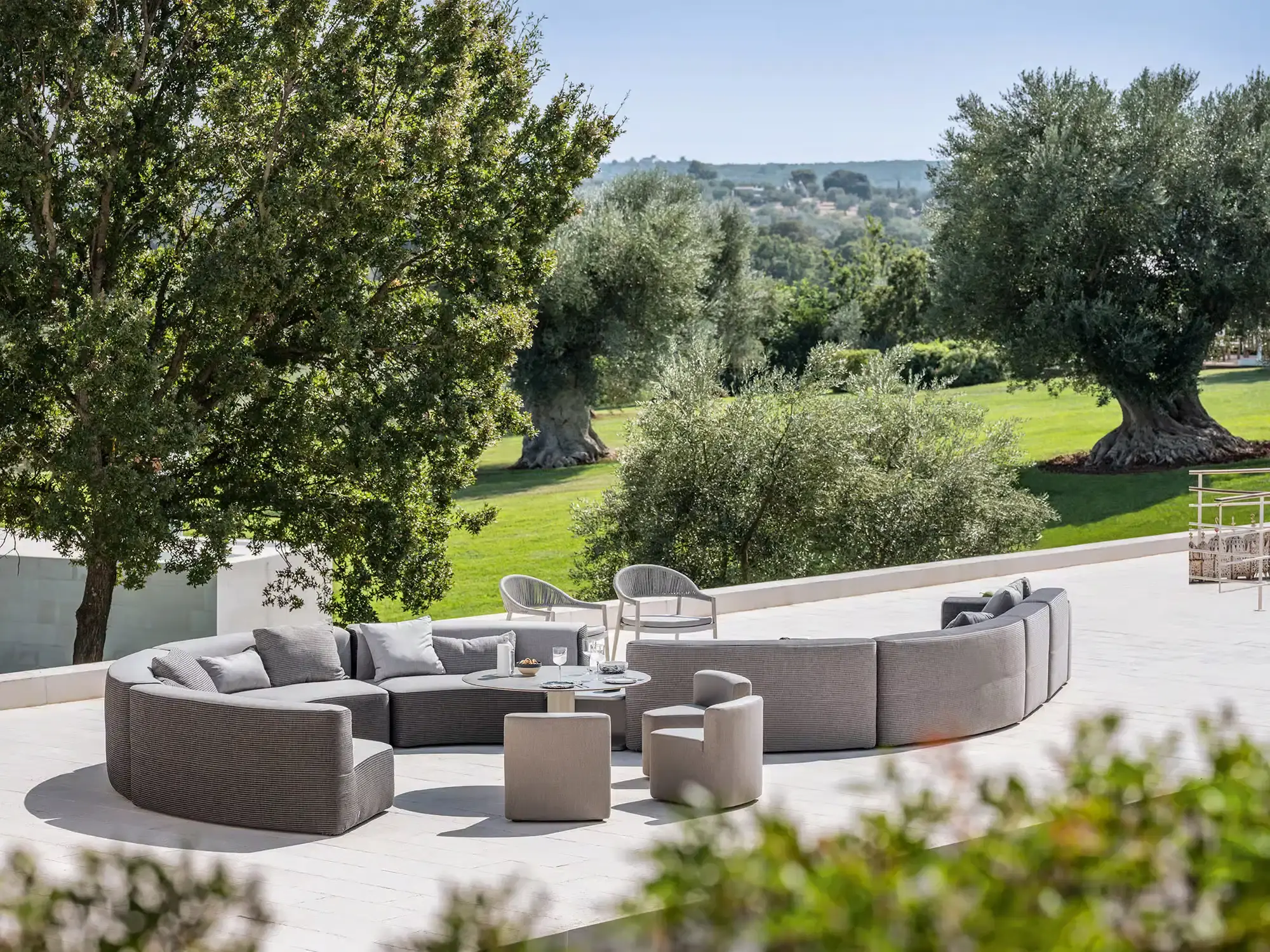 Große runde Outdoor-Lounge mit grauen Polstermöbeln, Sesseln und Couchtisch auf einer hellen Terrasse vor Olivenbäumen und weitem Blick ins Grüne.